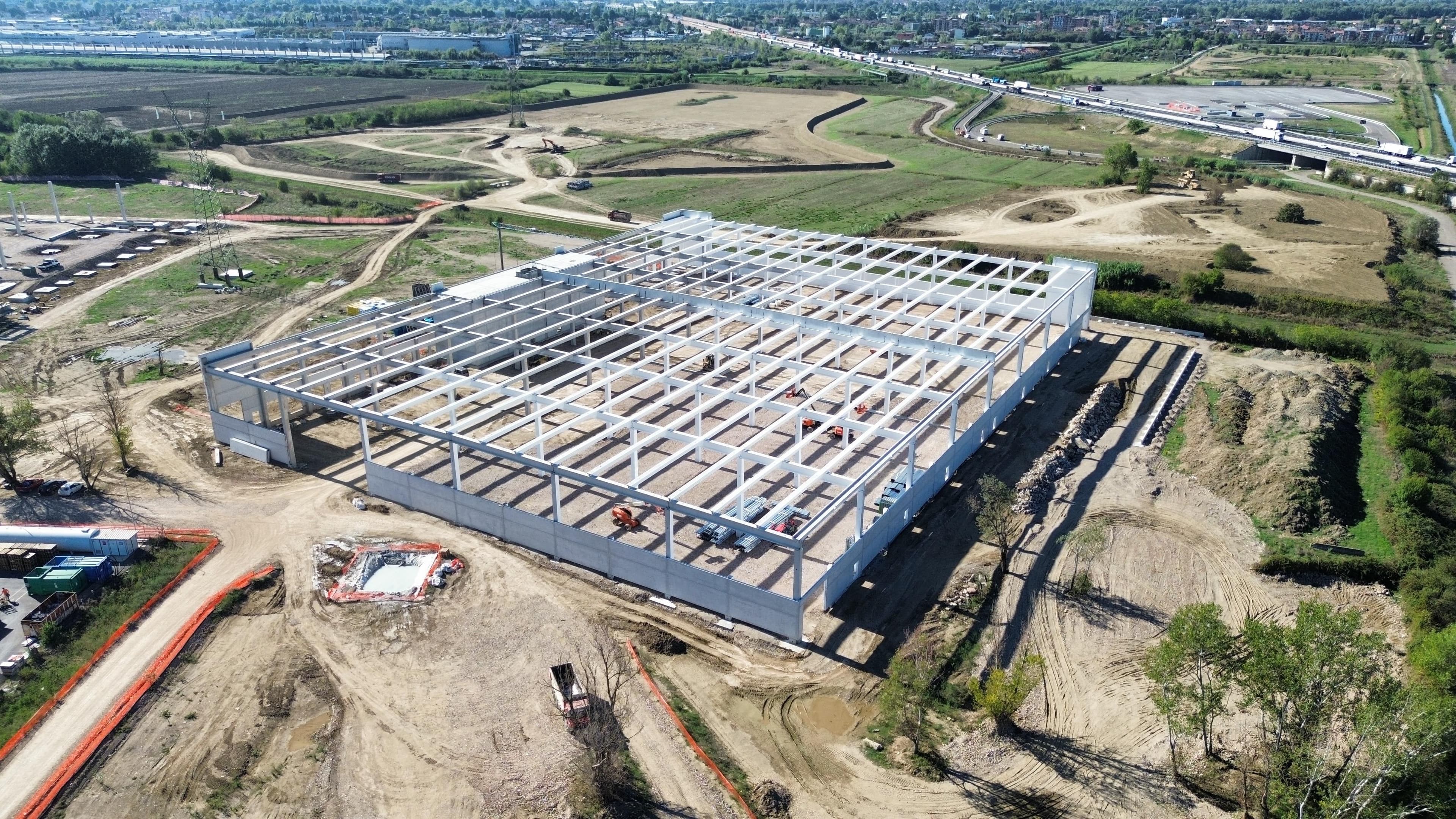 Aerial view of a large industrial building under construction, with a steel frame structure surrounded by dirt and undeveloped land.