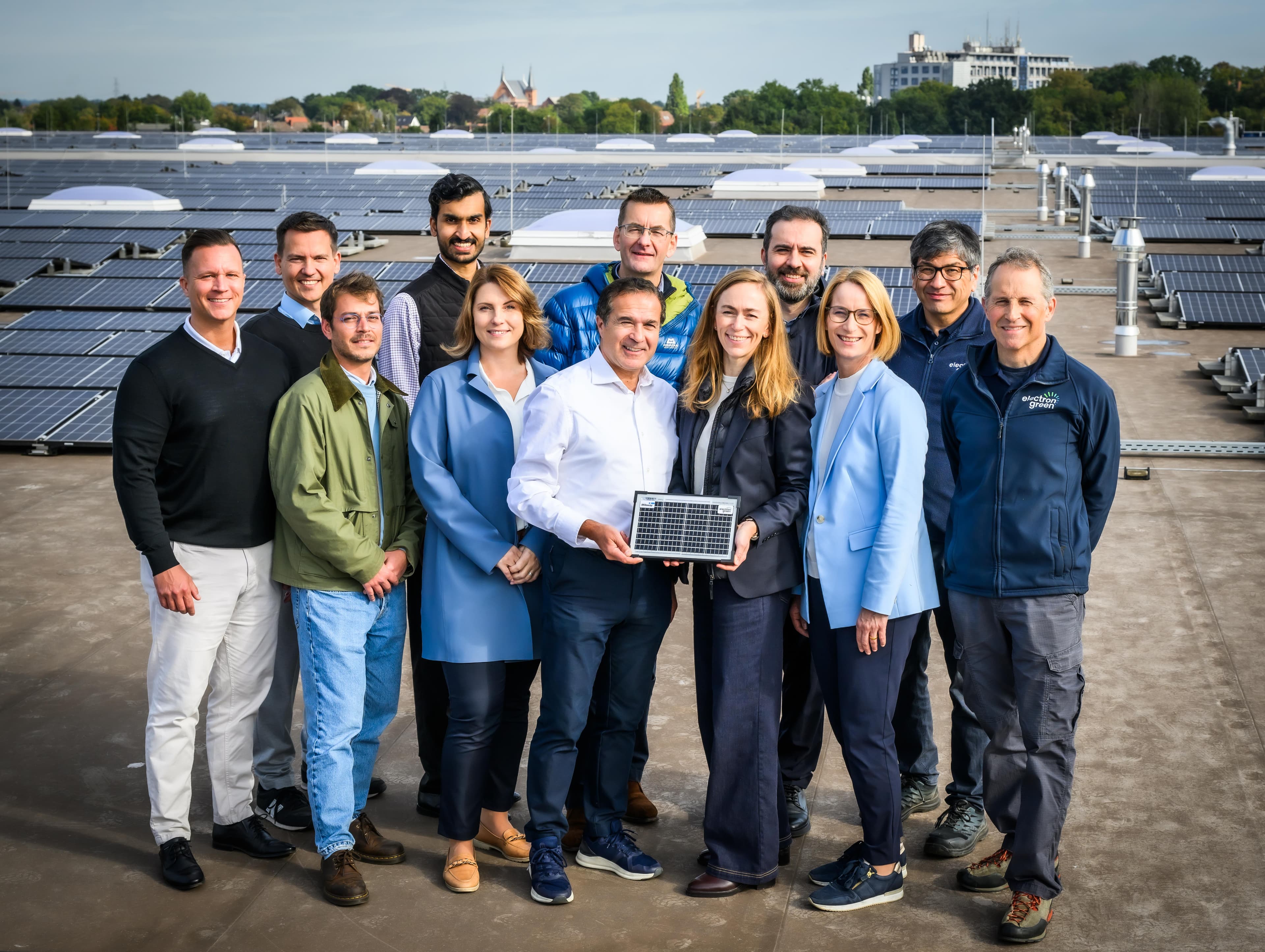 A group of twelve people stand on a rooftop with solar panels, with one person holding a small solar panel. Buildings and trees are visible in the background.