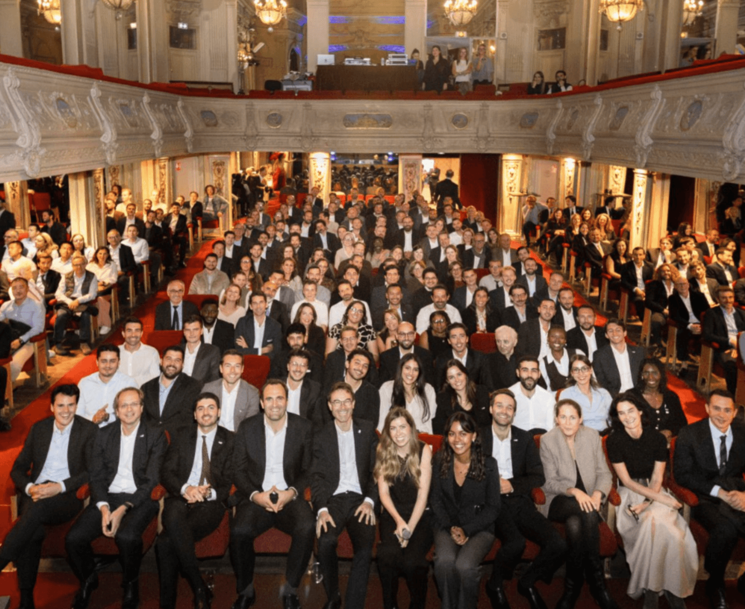 A large group of people dressed formally are seated and facing the camera in a grand theatre with ornate decor and red seats.
