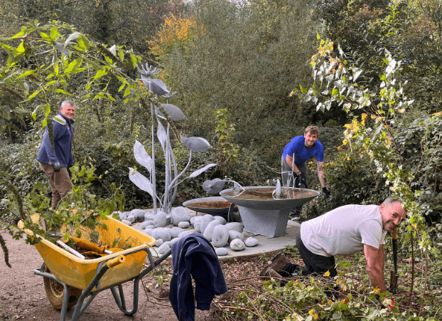 Three people are gardening near a metal sculpture surrounded by stones, with a wheelbarrow and foliage in a wooded outdoor area.