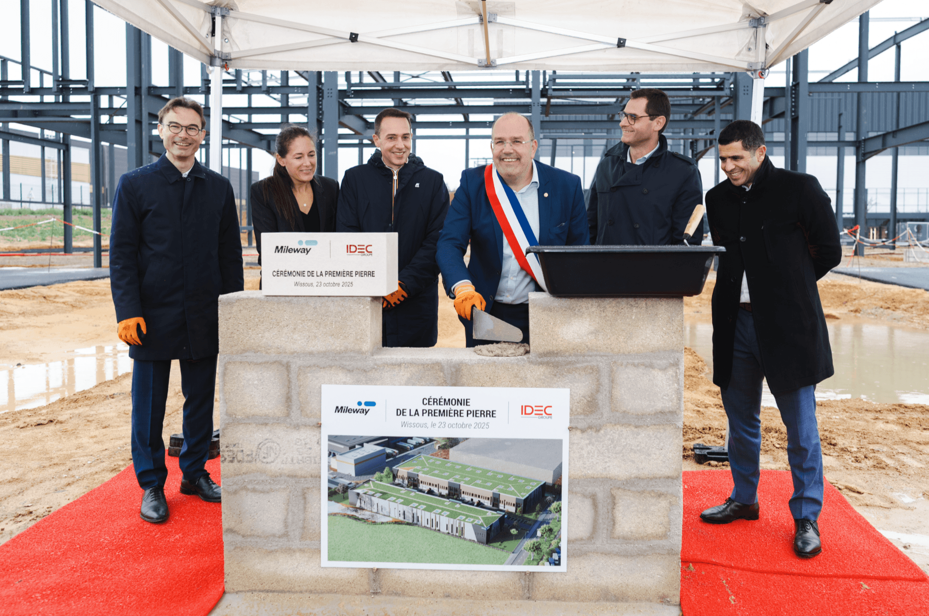 Six people in formal attire stand at a foundation stone ceremony site, with a plaque and architectural plan displayed, and steel framework construction in the background.