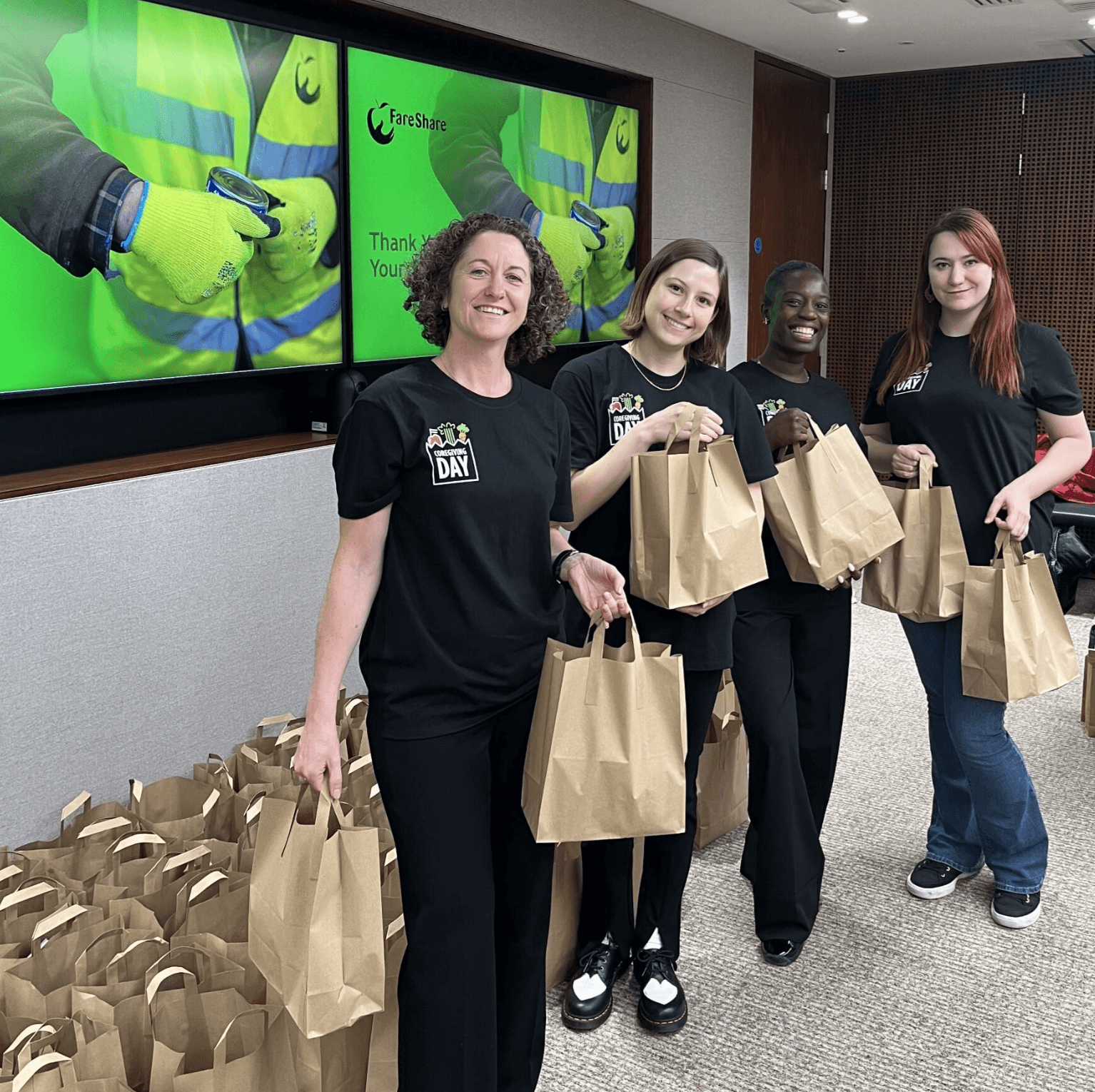 Four women wearing matching DAY shirts stand indoors holding brown paper bags, with more paper bags arranged on the floor around them; two screens with green backgrounds are visible behind them.
