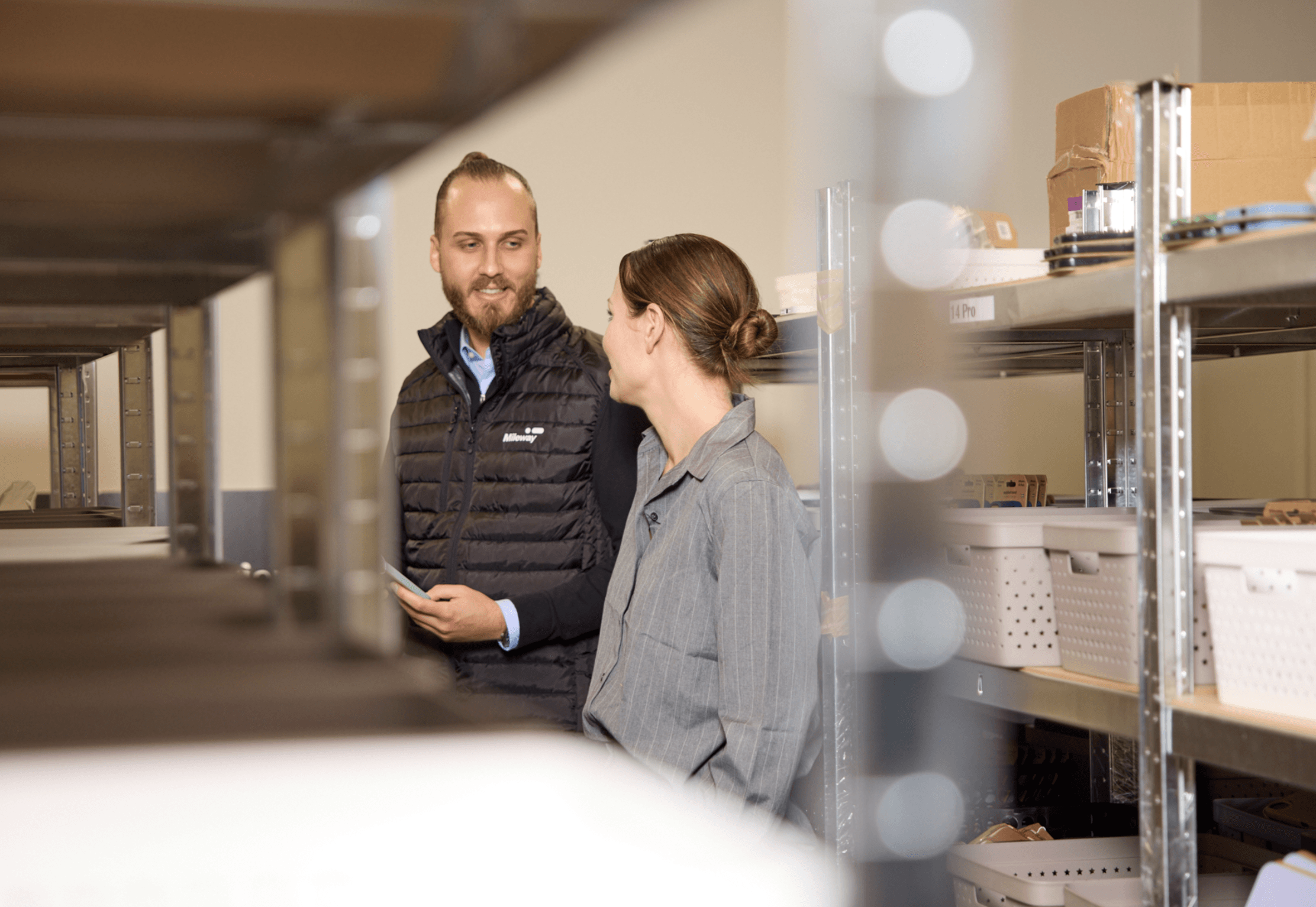 Two people stand and talk between metal shelves stocked with boxes and containers in a storage or warehouse setting.