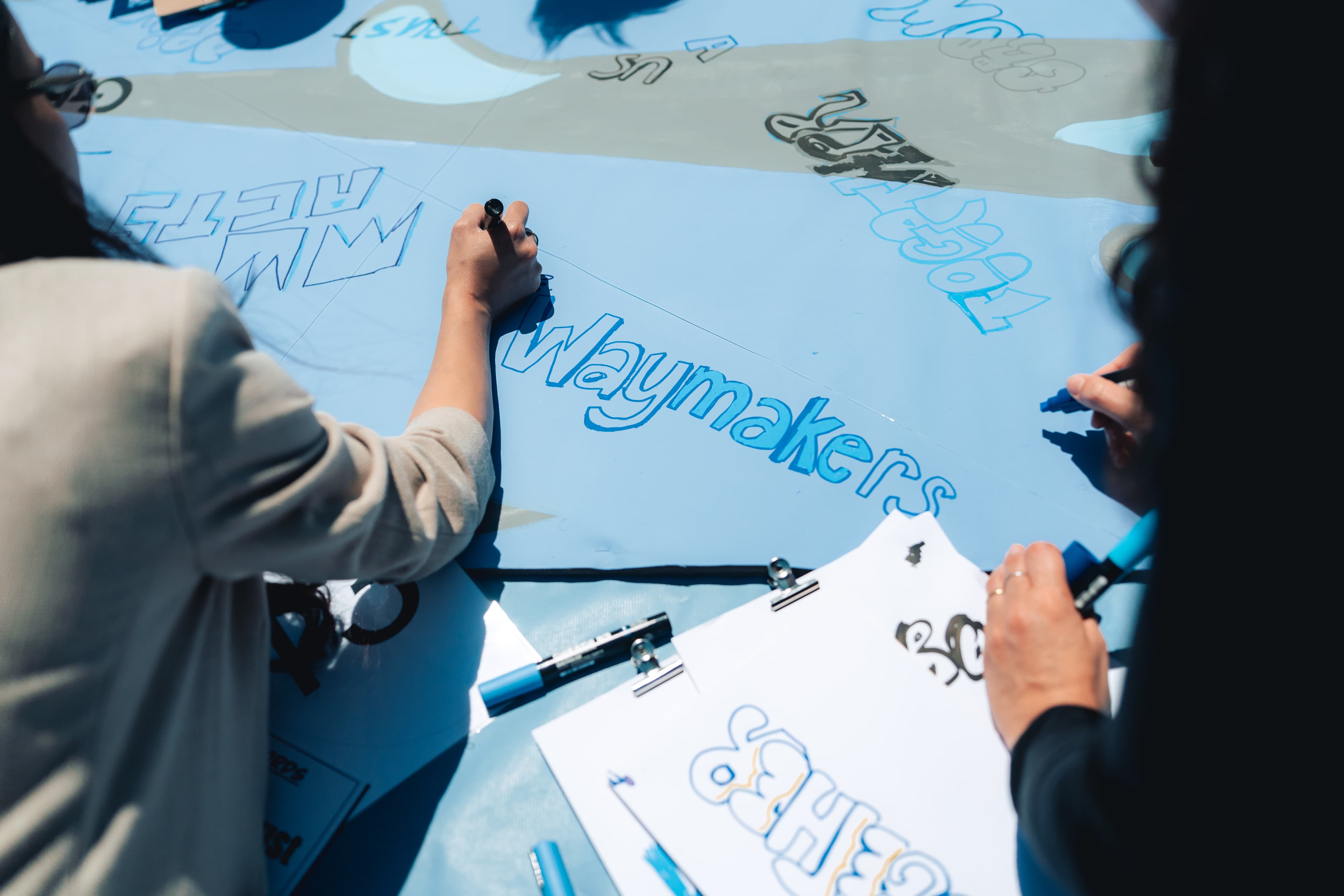 Close-up of people drawing and colouring words like Waymakers on a large blue poster, with clipboards holding paper and markers visible on the table.