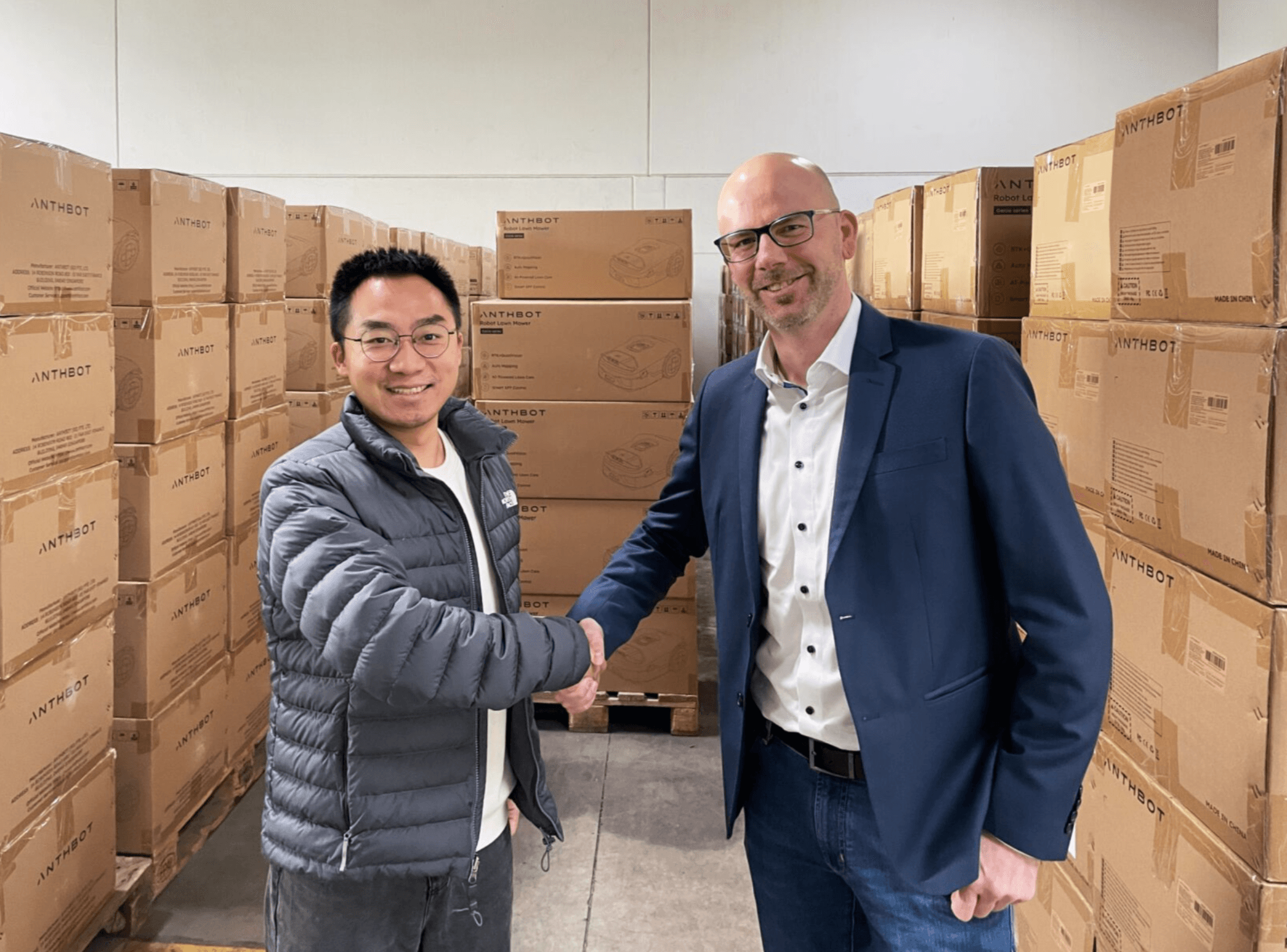 Two men shake hands in a warehouse, surrounded by stacked boxes labelled ANTH BOT.
