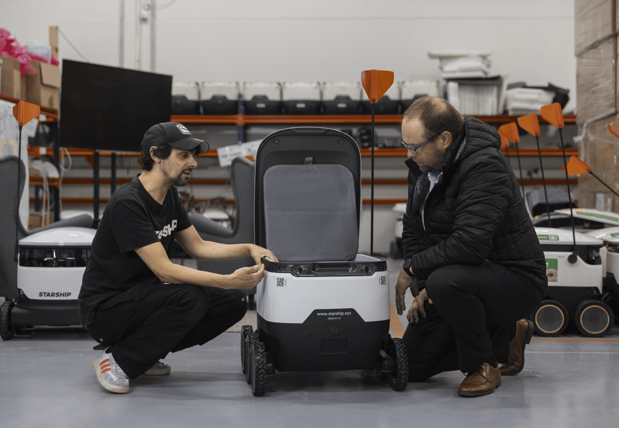 Two men kneel beside an open autonomous delivery robot in a warehouse, discussing its interior components.