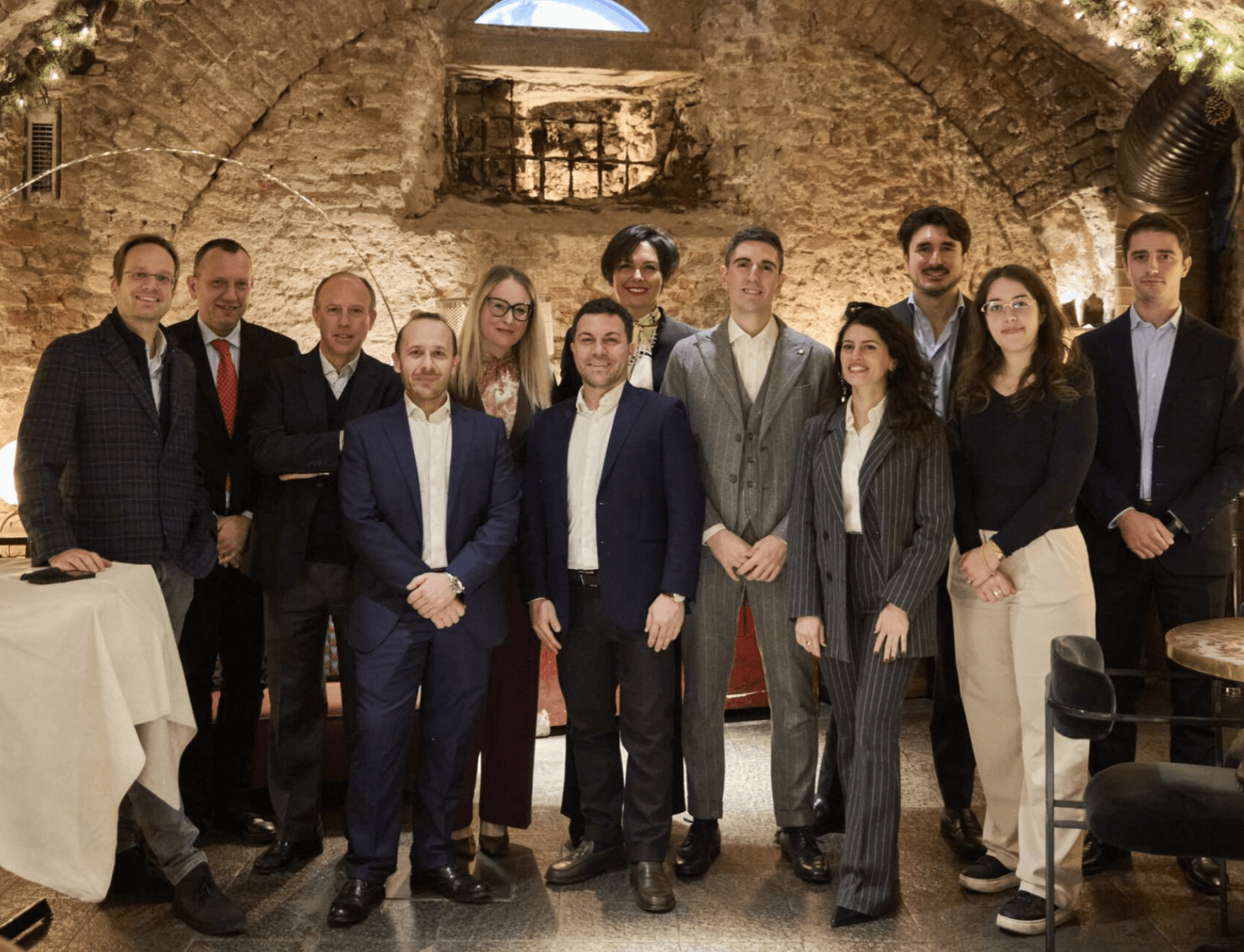 A group of twelve adults in business attire pose together in a rustic, brick-walled room with arched ceilings and soft lighting.