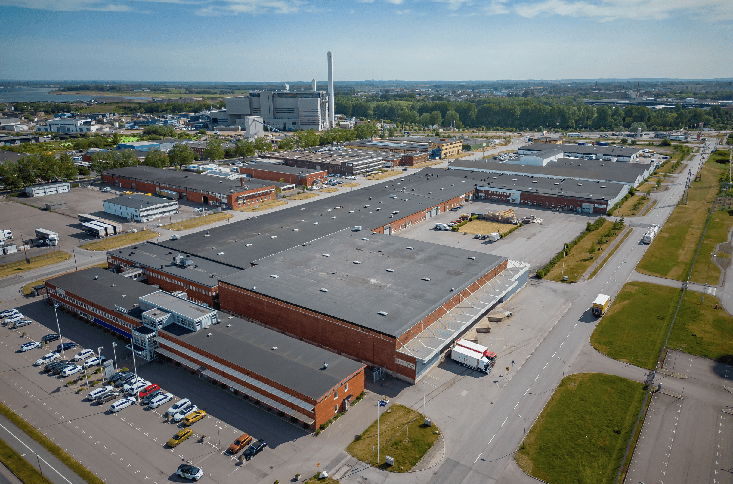 Aerial view of a large industrial complex with multiple warehouse buildings, lorries, parked cars, and roads, surrounded by greenery under a clear sky.