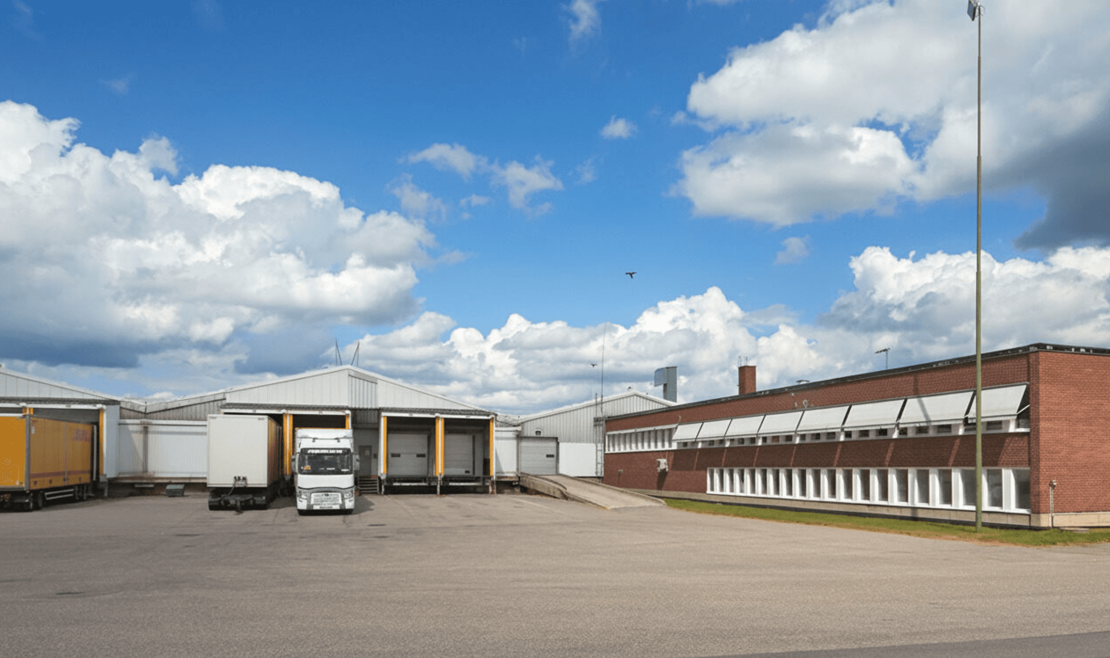A lorry is parked at loading bays outside a large industrial warehouse with brick and metal buildings under a partly cloudy sky.