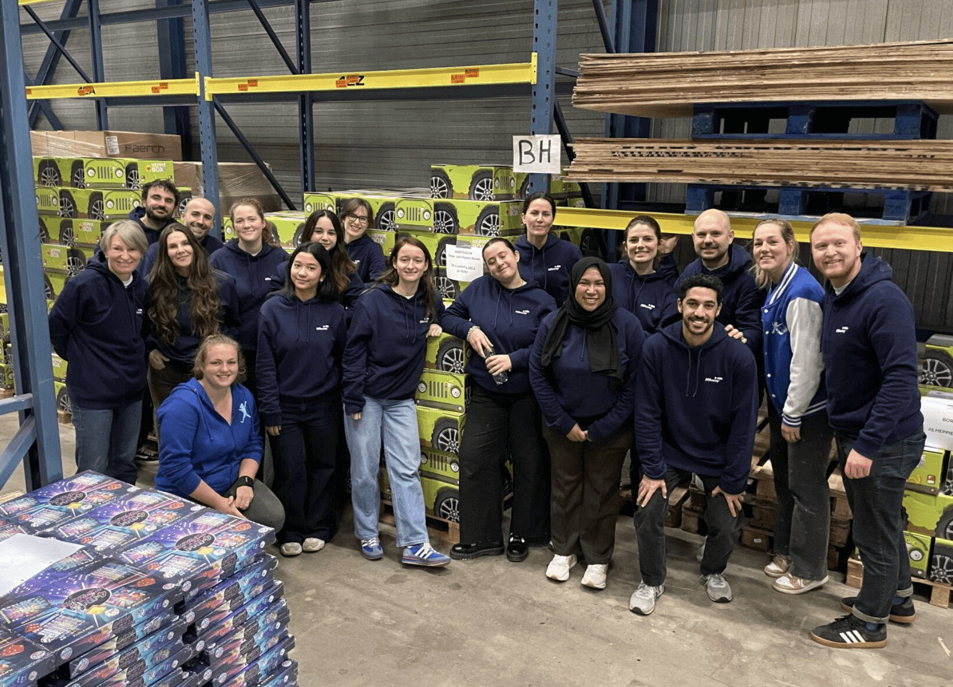 A group of people wearing blue and black hoodies pose together in a warehouse, standing in front of stacked boxes and wooden pallets.