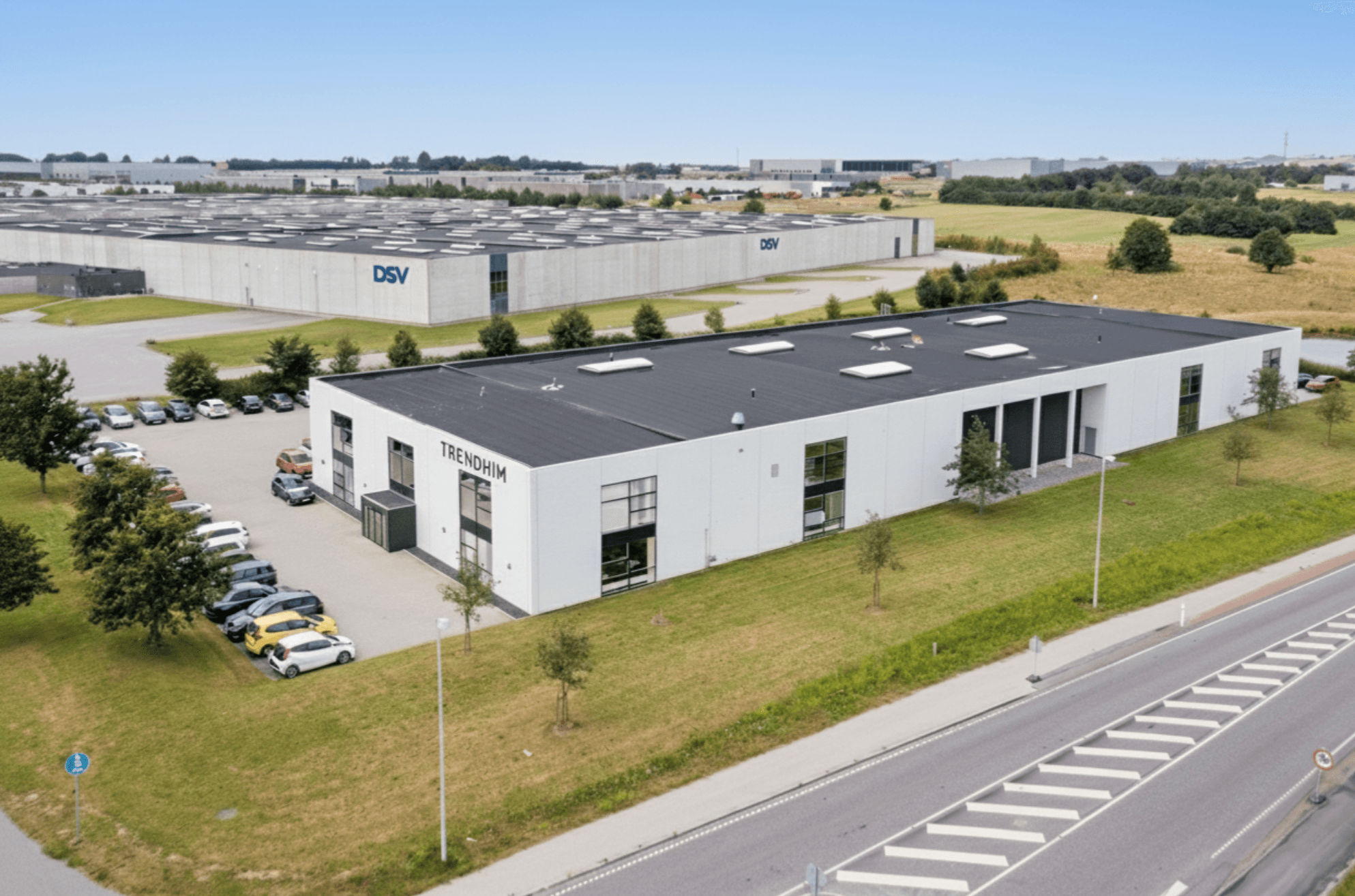 A large, modern white industrial building labelled Trekonim with a car park, grass lawn, and road in the foreground; similar buildings and fields in the background.