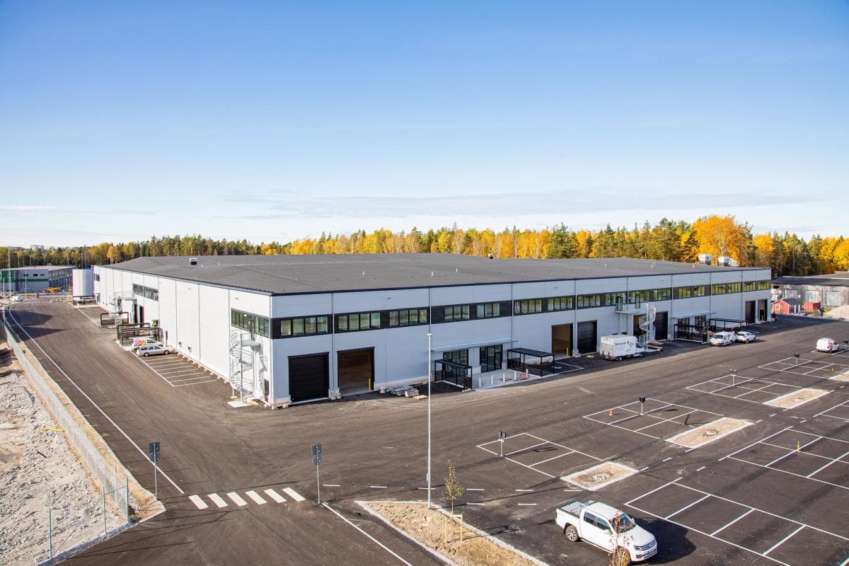 A large, white industrial warehouse with loading bays and several parked vehicles, surrounded by an empty car park and bordered by trees with autumn foliage.
