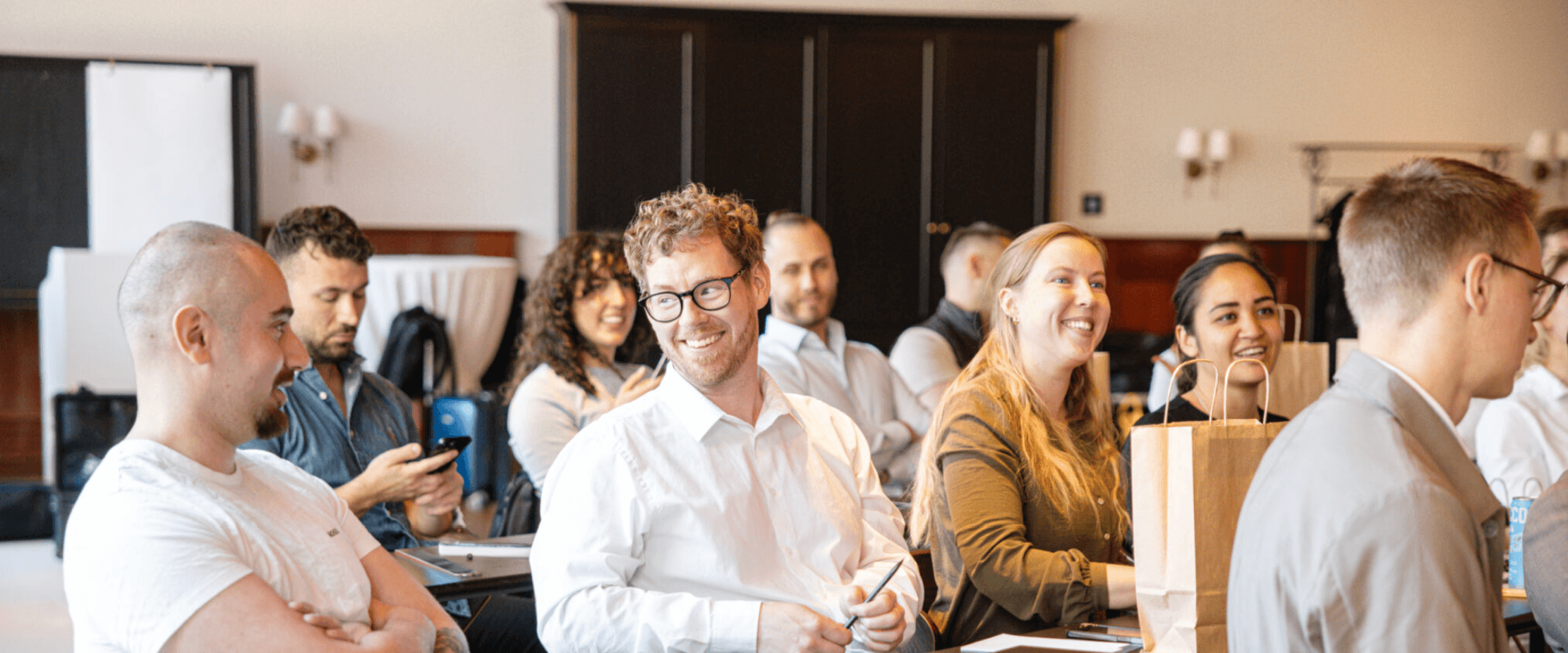 People sit at tables in a conference room, some smiling and engaged, with notebooks, pens, and gift bags on the tables.