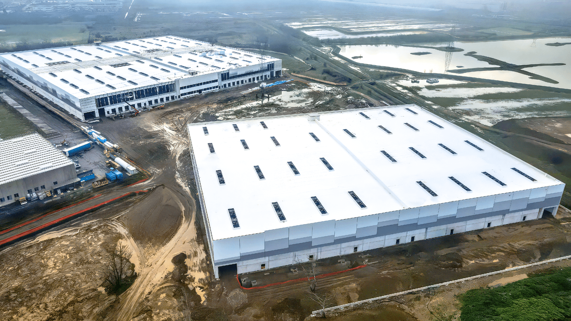Aerial view of two large, white-roofed industrial warehouse buildings surrounded by construction areas and patches of wet, undeveloped land.
