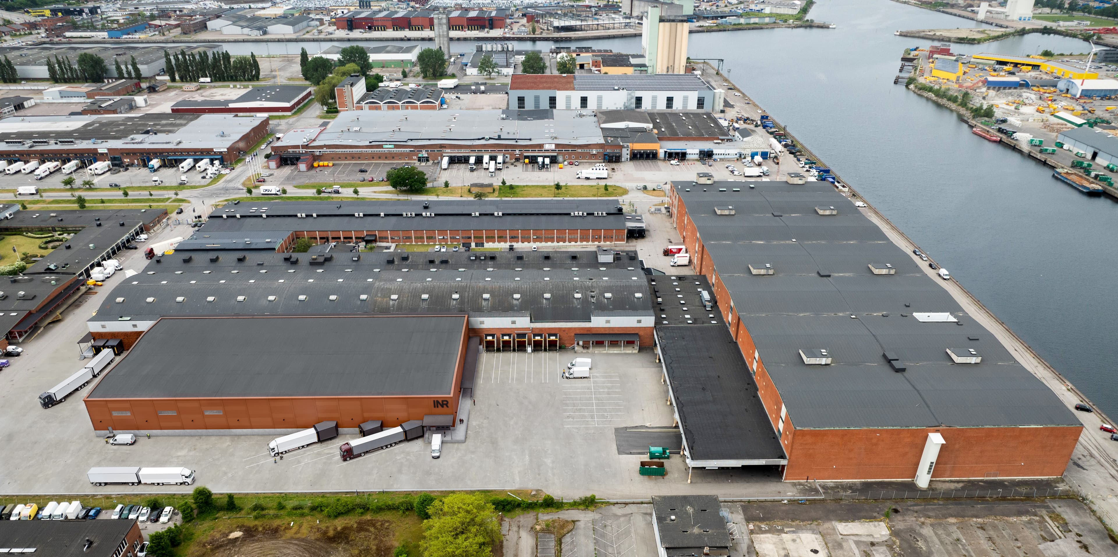 Aerial view of a large industrial warehouse complex with lorries parked outside, adjacent to a canal and surrounded by other industrial buildings.