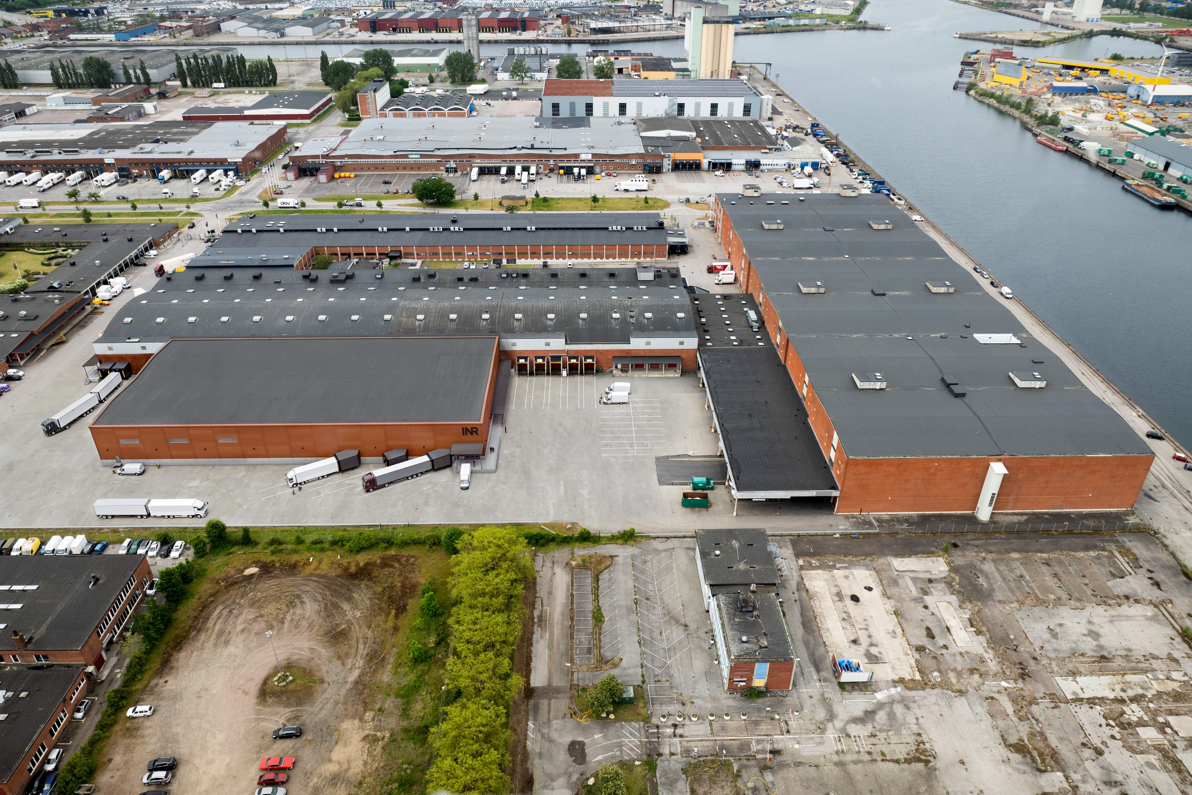 Flygfoto över ett stort industriellt lagerkomplex med lastkajer, parkerade lastbilar och en intilliggande vattenväg i ett stadsområde.