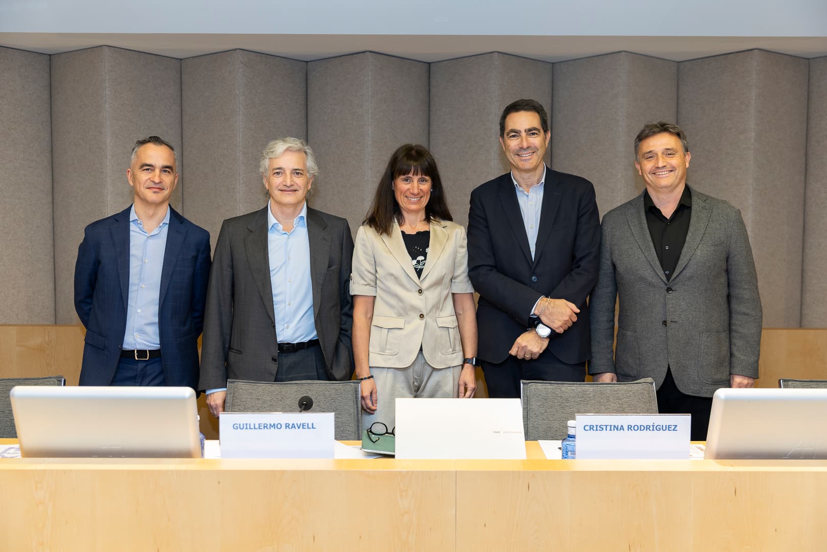 Several individuals in business attire stand behind a desk in a conference room, with nameplates and microphones on the table.