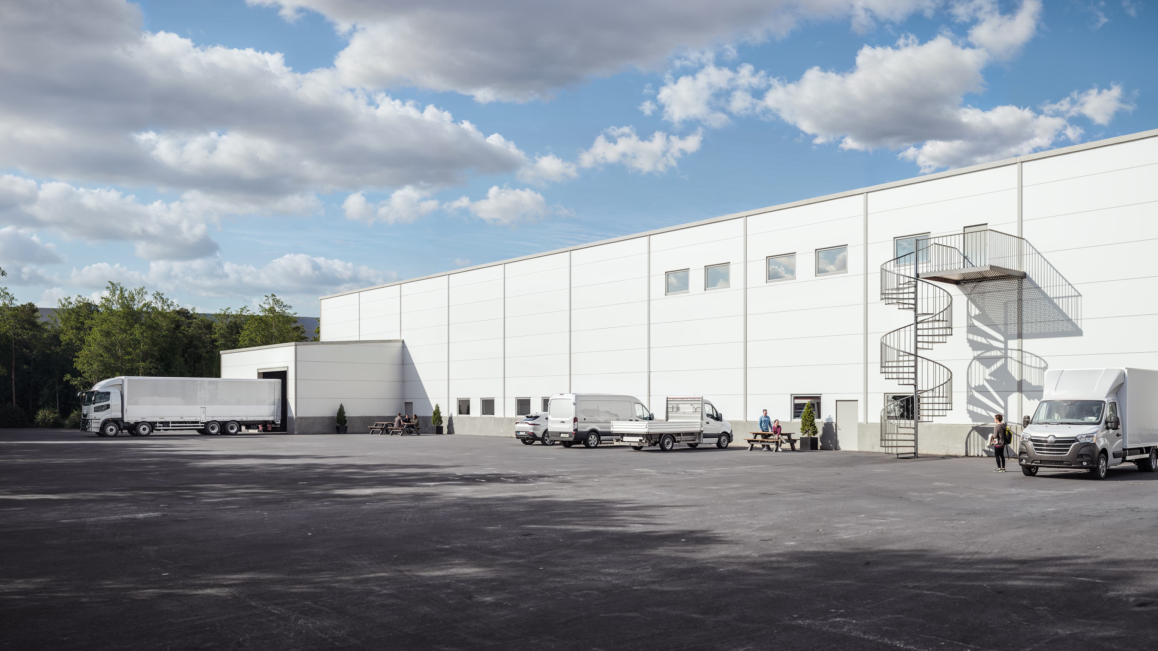 Large white warehouse with lorries and vans parked outside, individuals near the entrance, and a metal spiral staircase under clouds.