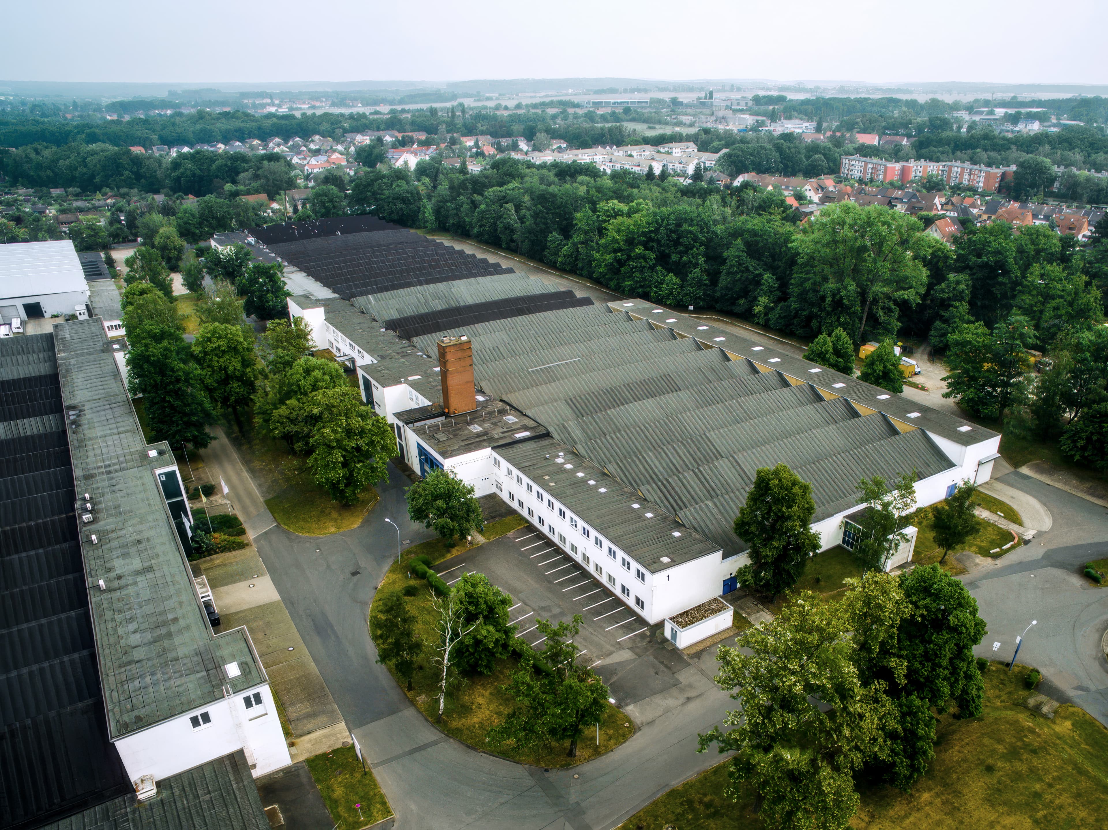 Aerial view of a large industrial warehouse complex bordered by trees, car parks, and a residential area in the background.