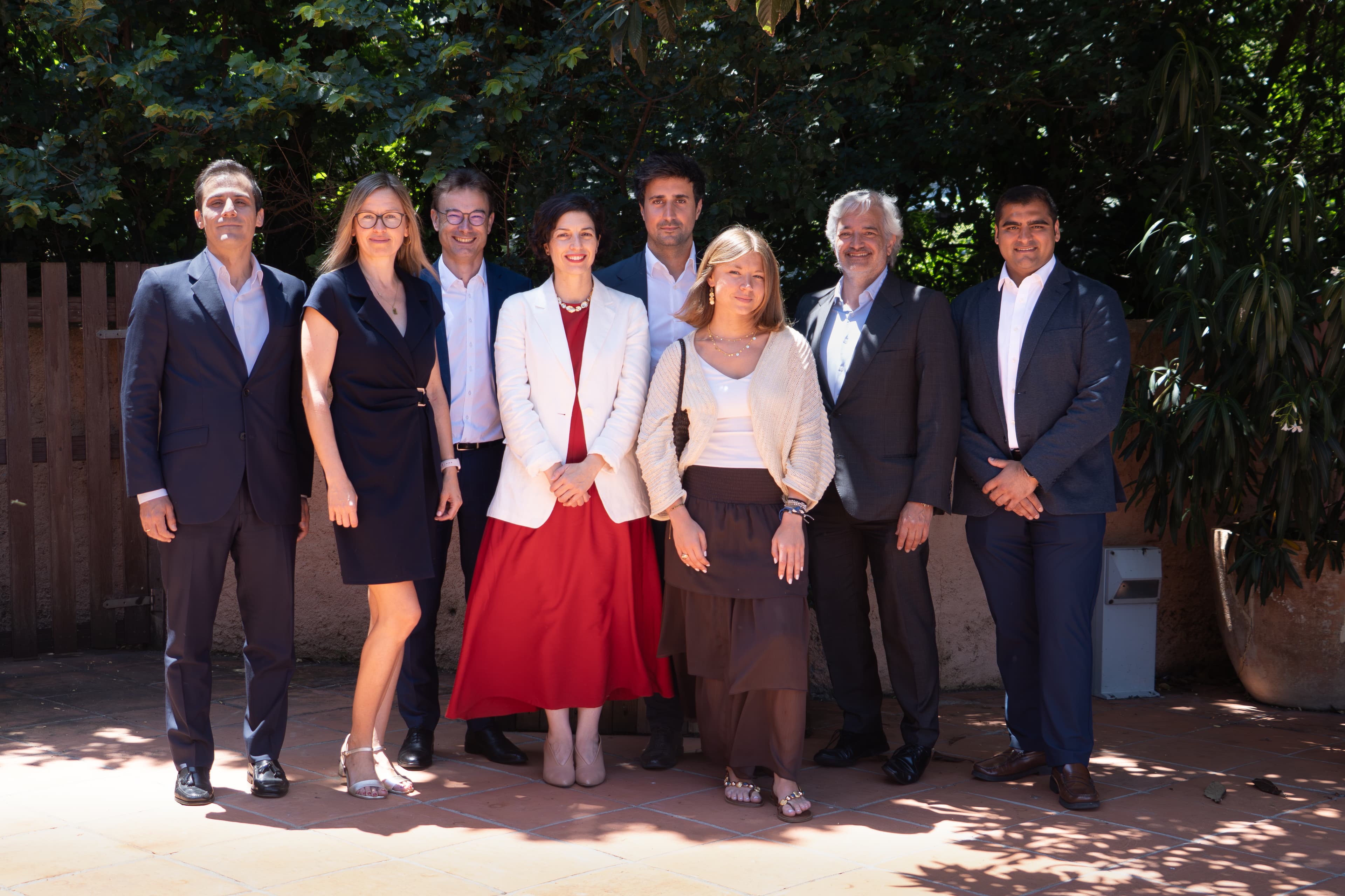 Eight people pose outdoors in business attire on a sunlit patio with trees and a wooden fence in the background.