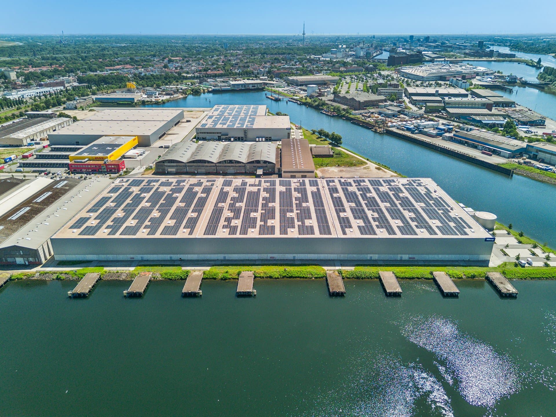 Vista aérea de un gran almacén industrial con paneles solares en el tejado, situado junto a un canal en una zona urbana.