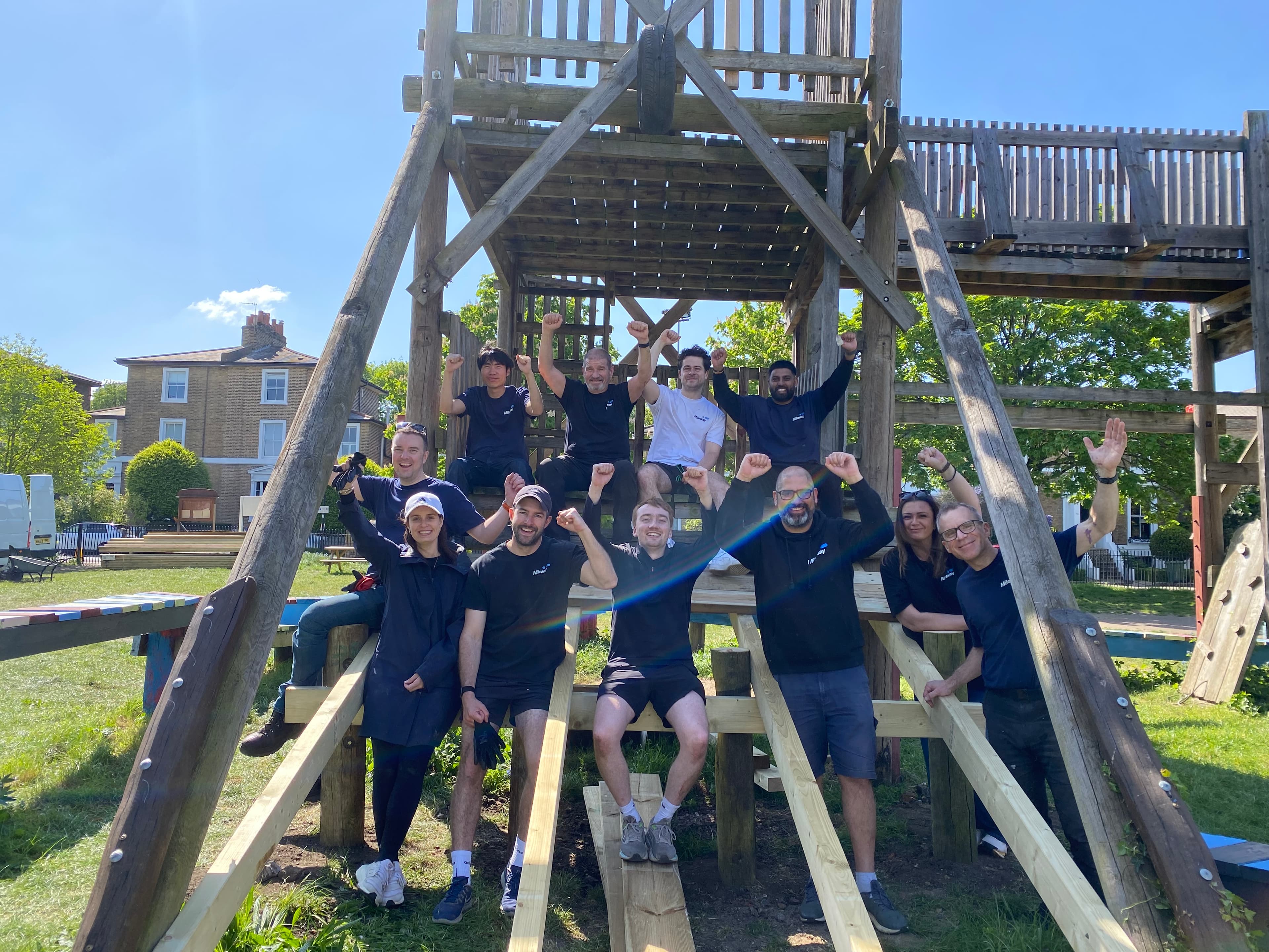 A group of people in outdoor clothing pose with raised arms and smiles on a wooden climbing frame in a sunny park.