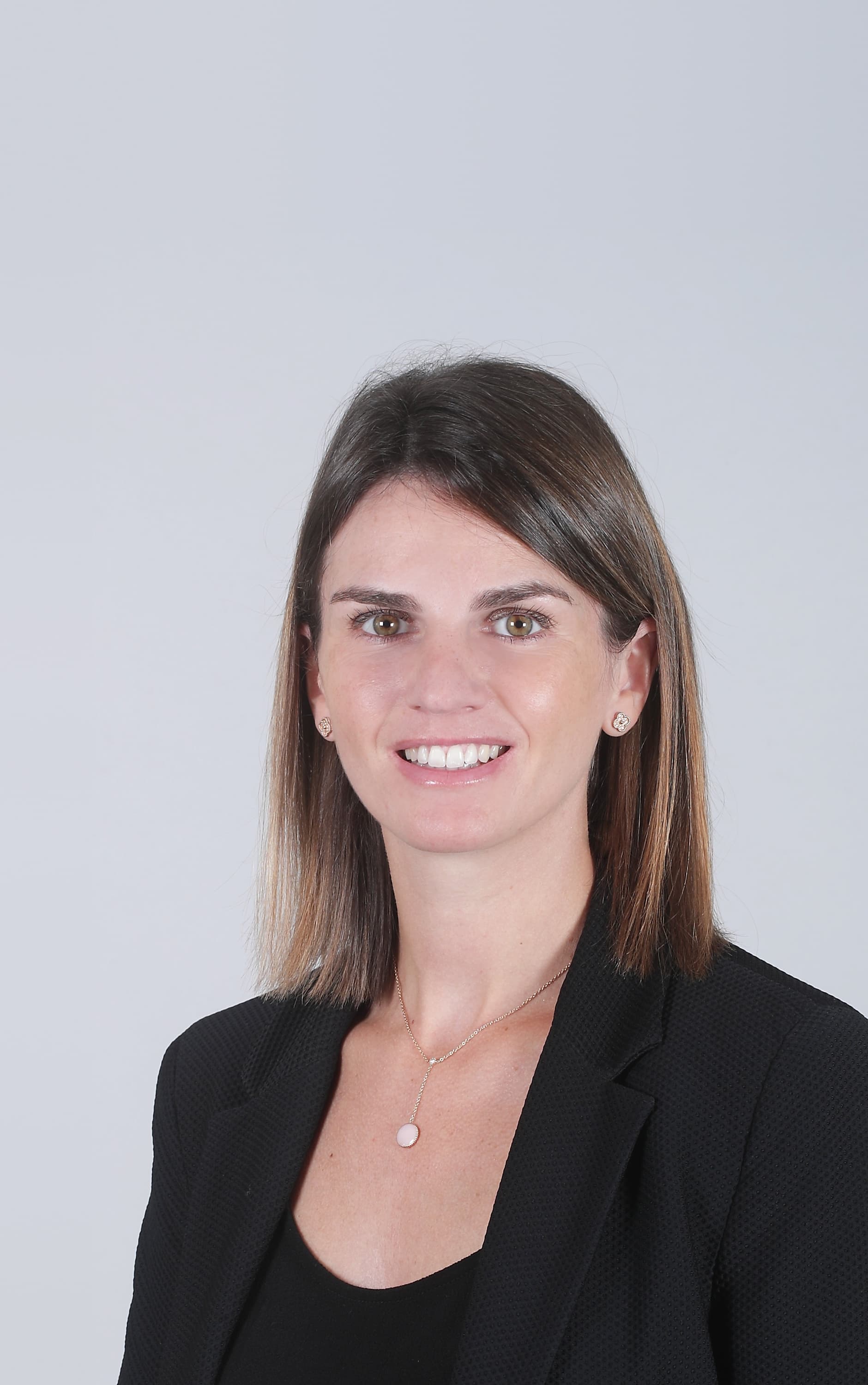 A person in a black blazer smiles at the camera against a plain light grey background.